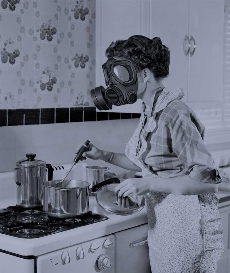 A black and white photo of a women wearing a gas mask and cooking over a gas stove.